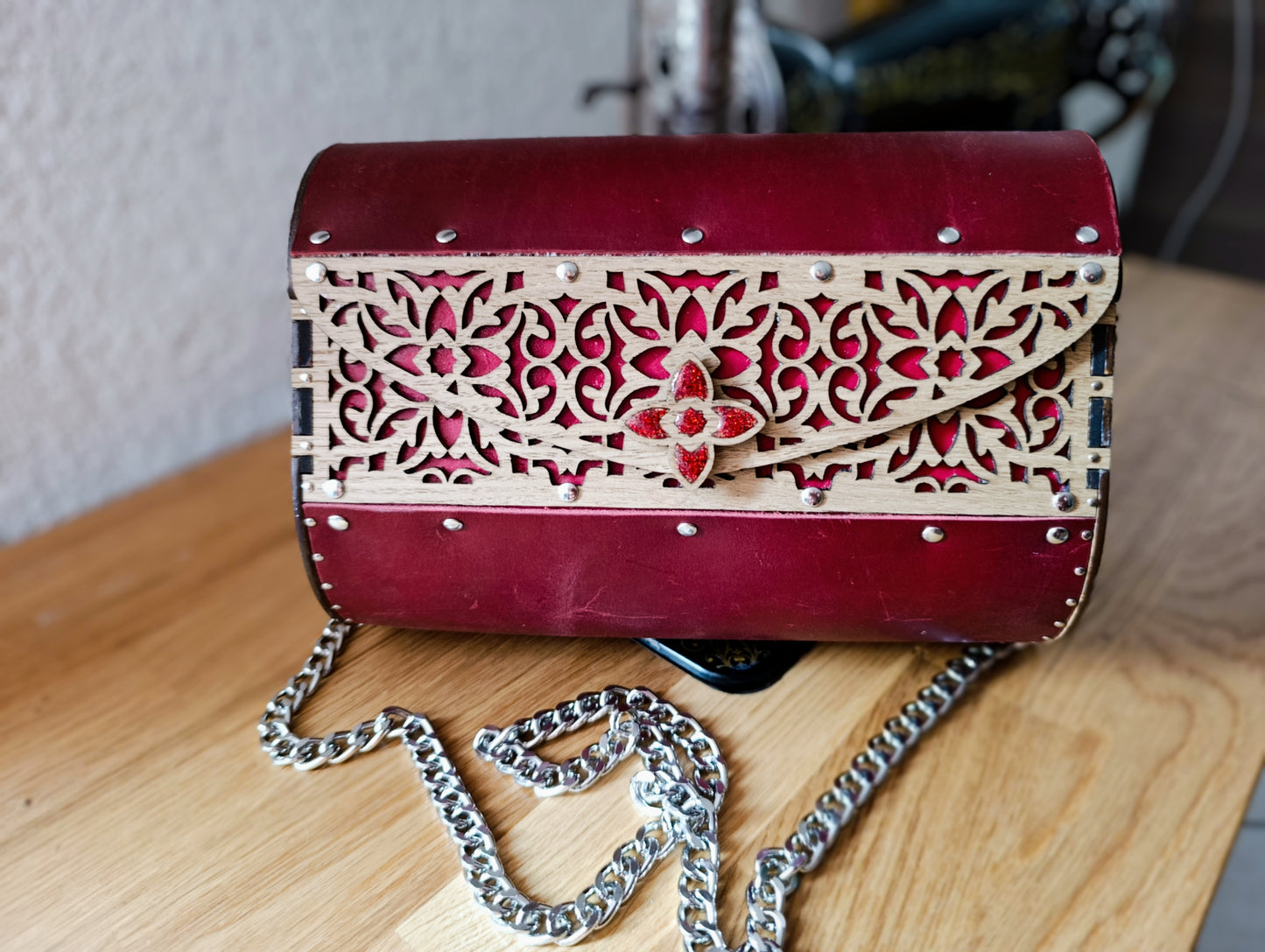 Pochette en cuir rouge foncé et bois ajouré gravé, vue de face avec fermoir en résine rouge pailletée, posée sur une table en bois.