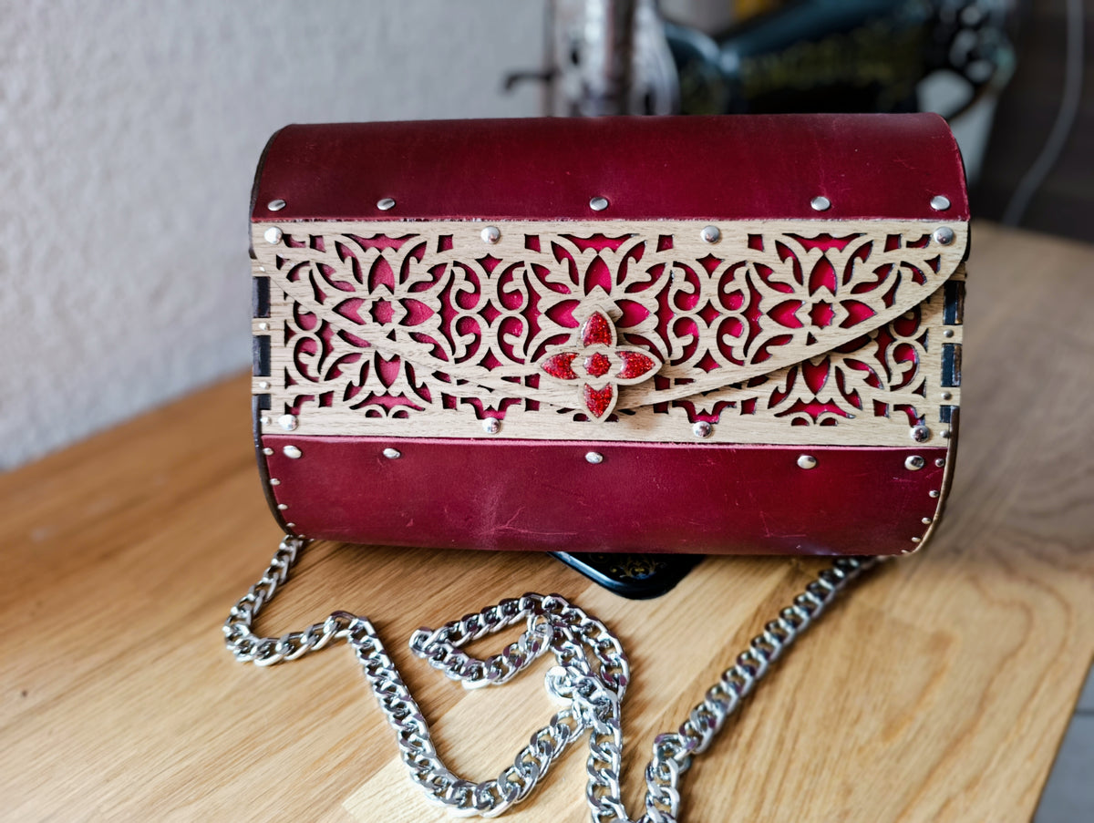 Pochette en cuir rouge foncé et bois ajouré gravé, vue de face avec fermoir en résine rouge pailletée, posée sur une table en bois.